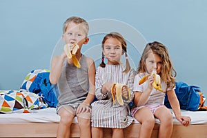 Three kids in sleeping clothes sitting on unmade bed, eating bananas