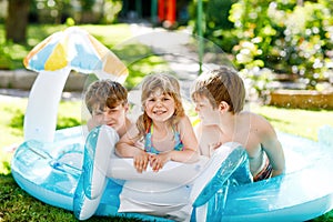 Three kids, two boys and toddler girl splash in an outdoors swimming pool in summer. Happy children, brothers and sister playing