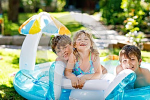 Three kids, two boys and toddler girl splash in an outdoors swimming pool in summer. Happy children, brothers and sister