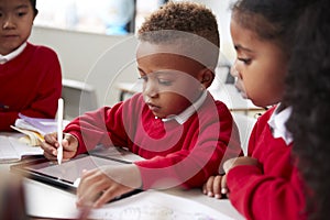 Three kindergarten school kids sitting at desk in a classroom using a tablet computer and stylus together, selective focus