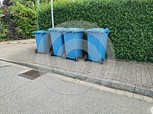 Three large and one small blue plastic garbage cans stands on the side of the road in front of a green hedge ready for collection