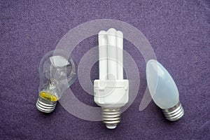 Three light bulbs. A white energy-saving fluorescent bulb with four tubes, an oblong incandescent bulb with gray glass, a round or