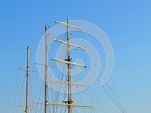 Three masts of a sailing ship with folded up sails and shrouds against a blue summer sky