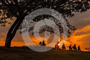 Three men dressed in cowboy garb, with horses and guns. A cowboy riding a horse in the sunset is silhouetted in black