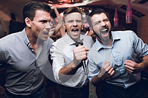 Three men sing at a karaoke club. Young people have fun in a nightclub. They are very cheerful and they smile.