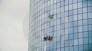 Three men workers hanging on ropes and cleaning the exterior glass windows of a business skyscraper after rain - autumn