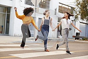 Three millennial hip girlfriends holding hands and laughing as they run across a pedestrian crossing