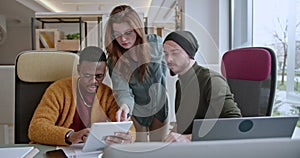 Three, mixed race colleagues engaged in a business discussion, examining data on a tablet and laptop in a well-lit