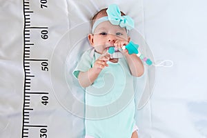 A three month baby girl in mint green clothes lying on a bed on which a measuring ruler for growth is drawn. Teething tool in