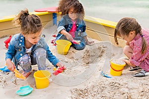 three multiethnic little children playing with plastic scoops and buckets in sandbox