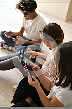 Three multiethnic little kids sit indoor using digital gadgets