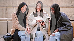 three multiracial women friends smiling using a smartphone looking for clothes on internet. A group of diverse girls