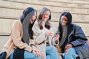 three multiracial women friends smiling using a smartphone looking for clothes on internet. A group of diverse girls