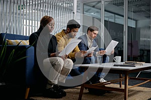 Three multiracial young adults in businesswear sitting on sofa and reading documents in office