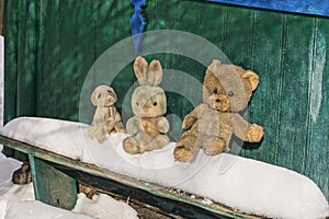 Three old plush toy animals stand on a wooden bench in white snow