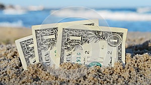 Three one dollar banknotes dug halfway into sand on beach near sea on sunny day