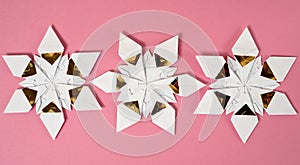 Three origami star snowflakes arranged in a row in white and gold foil paper, on a pink paper background