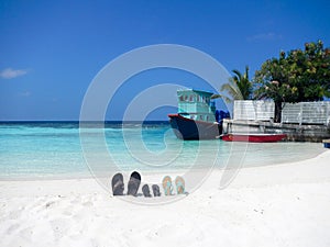 Three pairs of flip flops sandals on a Maldive beach propped up in the sand with an old fishing boat at a quay behind and palm tre