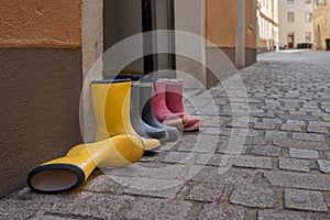 Three pairs of rubber boots for children in different colours standing on the street next to front door of house