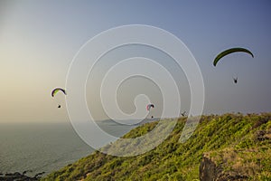 Three paragliders on colorful parachutes fly over the sea and the green hill against a clean blue sky