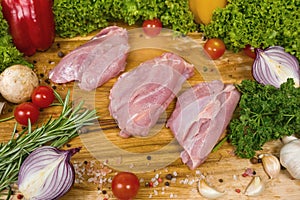 Three pieces of turkey meat on a cutting wooden board with cherry tomatoes, lettuce leaves, garlic, chilli, rosemary