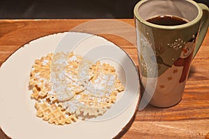 Three pizzelles covered with powdered sugar, on a round, white. plate and a Christmas mug of hot coffee