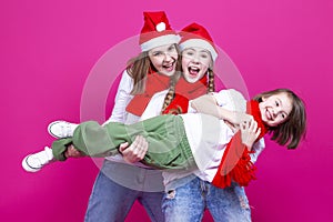 Three Positive Caucasian Girls With Santa Hats Having Fun While Lifting Teenage Girl Holding Hands Closed Against Trendy Pink