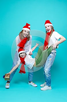 Three Positive Caucasian Girls With Santa Hats Having Fun While Lifting Teenage Girl Holding Wrapped Gift Box Against Trendy Green