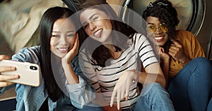 Three pretty mixed-races friendly girls at washing machines in washhouse smiling to smartphone camera, taking selfie