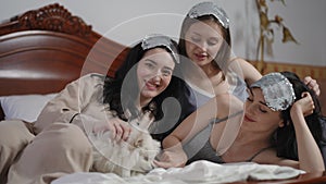 Three pretty young women, with silver sleep masks on their heads, and a white Spitz dog, posing while lying on the bed