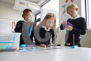 Three primary school children working together with toy construction blocks in a classroom, the girl reading instructions from a b