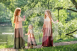 Three red-haired sisters in long linen dresses blow bubbles in the park on sunny summer day