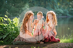 Three red-haired sisters in long linen dresses blow bubbles in the park on sunny summer day