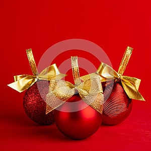 Three red shiny christmas balls with gold shimmer bow on bright deep red background, square, closeup. New year background.