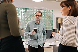 Three relaxed multiethnic coworkers standing in office, drinking coffee and talking cheerfully in workspace during break