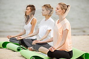 Three relaxed young girls sit in the lotus positions with closing eyes doing yoga on mats on sandy beach on a warm day