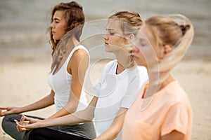 Three relaxed young girls sit in the lotus positions with closing eyes doing yoga on mats on sandy beach on a warm day