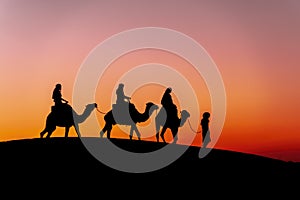 Three Riders And Their Handler Travel Through The Saharan Desert On Their Camels In Morocco