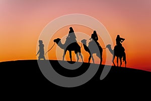 Three Riders And Their Handler Travel Through The Saharan Desert On Their Camels In Morocco