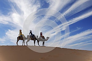 Three Riders And Their Handler Travel Through The Saharan Desert On Their Camels In Morocco