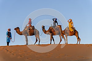 Three Riders And Their Handler Travel Through The Saharan Desert On Their Camels In Morocco