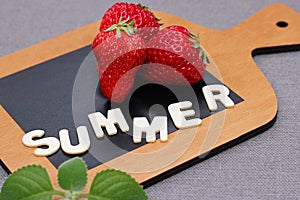 Three ripe red fresh strawberry on a wooden chopping board next to the word SUMMER in wooden letters. mint leaf