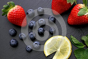 Strawberries, lemon slices, mint leaves and blueberries on black slate background