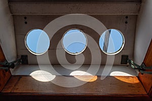 Three round portholes seen from inside a ship looking out on the ship`s rigging and blue sky