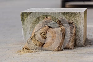 Three sandbags, one ripped open, for damming leaning on stone bench in street, ready for use against flood