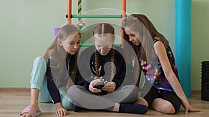 Three school girl using mobile phone in gym class at break. Teenager girl friends looking smartphone sitting on floor in