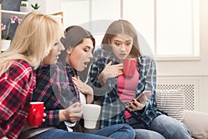 Three shocked female friends with coffee and smartphone at home