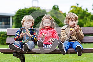 Three siblings sitting on bench and eating chocolate.