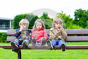 Three siblings sitting on bench and eating chocolate.