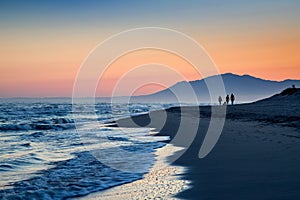 Three silhouettes of people stroll along the beach of Cabopino, Marbella, Malaga during sunset